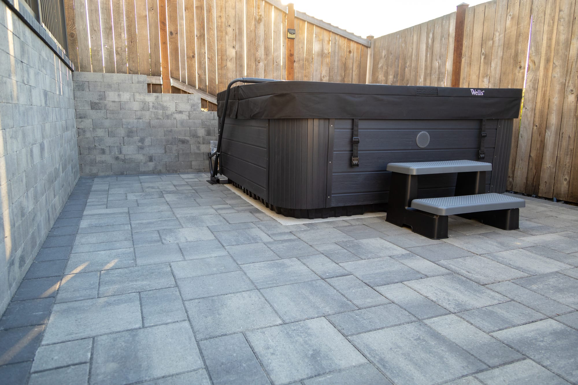 The hot tub on the lower paver patio, boxed in by grey stone retaining walls and a cedar fence